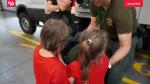 Dwoje dzieci odwróconych tyłem stoi przy funkcjonariuszu Straży Granicznej, który pokazuje im hełm.  Dzieci ubrane są ubrane w czerwone koszulki.