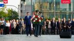 Zdjęcie z uroczystości obchodów 68 rocznicy Poznańskiego Czerwca. Funkcjonariusz ubrany w mundur niesie wieniec kwiatów, za nim idzie kolejny funkcjonariusz  w mundurze i mężczyzna w garniturze. Za nimi stoi tłum osób. 
